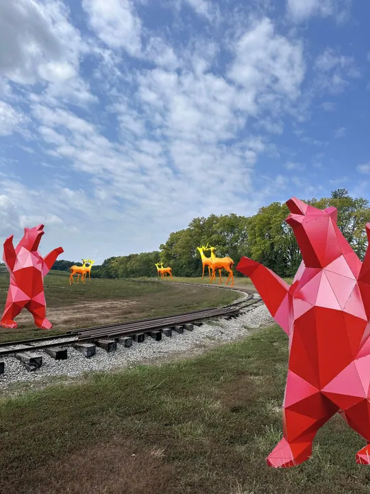 Faceted red bear sculptures standing beside a railway with golden deer in the distance